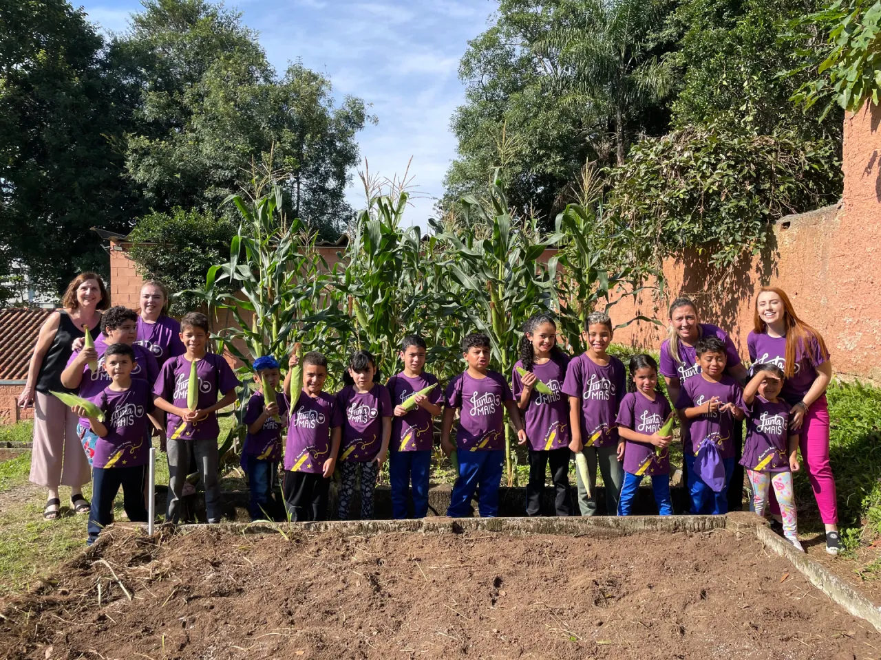 Grupo de crianças e educadoras da Junta Mais segurando espigas de milho na horta, em Curitiba